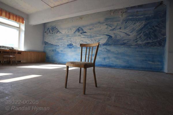 Empty chair and grand mural depicting nearby glacier are lonely reminders of better days at the Russian coal mining town of Pyramiden, abandoned in 1998; Isfjorden, Svalbard, Norway.