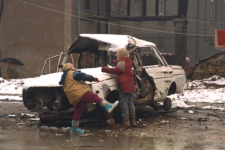 Children having a snowball fight amid the debris of war contrasted with news photos of children's bodies in the morgue. The childrens' deaths helped build pressure in the U.S. Congress for a unilateral lifting of the arms embargo. The dead children were victims of Serbian attacks on Sarajevo.