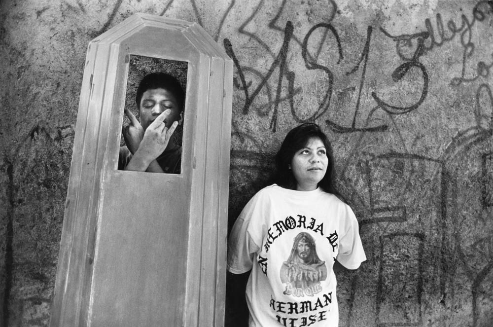 Sonia, a young Los Angeles girt, wears a memorial T-shirt from her brother Ulises’ gang funeral. A local Mara Salvatrucha gang member raises the lid of the coffin he was sanding to strike a pose of respect to the memory of his dead homeboy. Photo by APF Fellow Donna Decesare