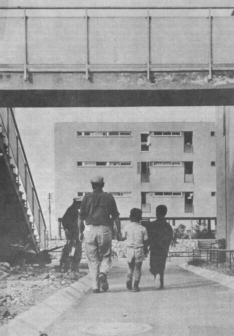 A small pedestrian path leads between two buildings and under a second story pedestrian bridge toward a larger pedestrian "street" in the model neighborhood at Kiryat Gato (Ministry of Housing photo)